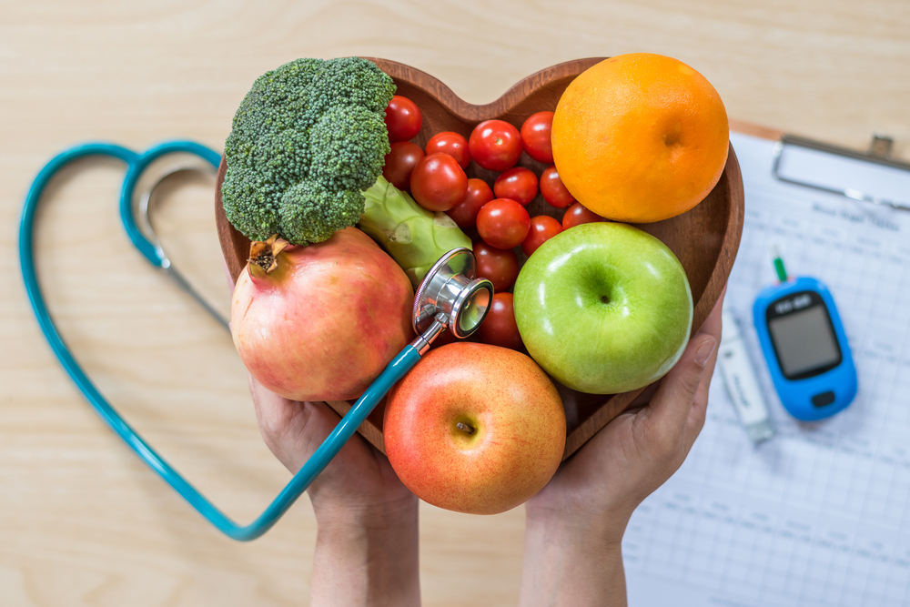 stethoscope in heart-shaped bowl with fruits and veggies over blood sugar monitor and chart